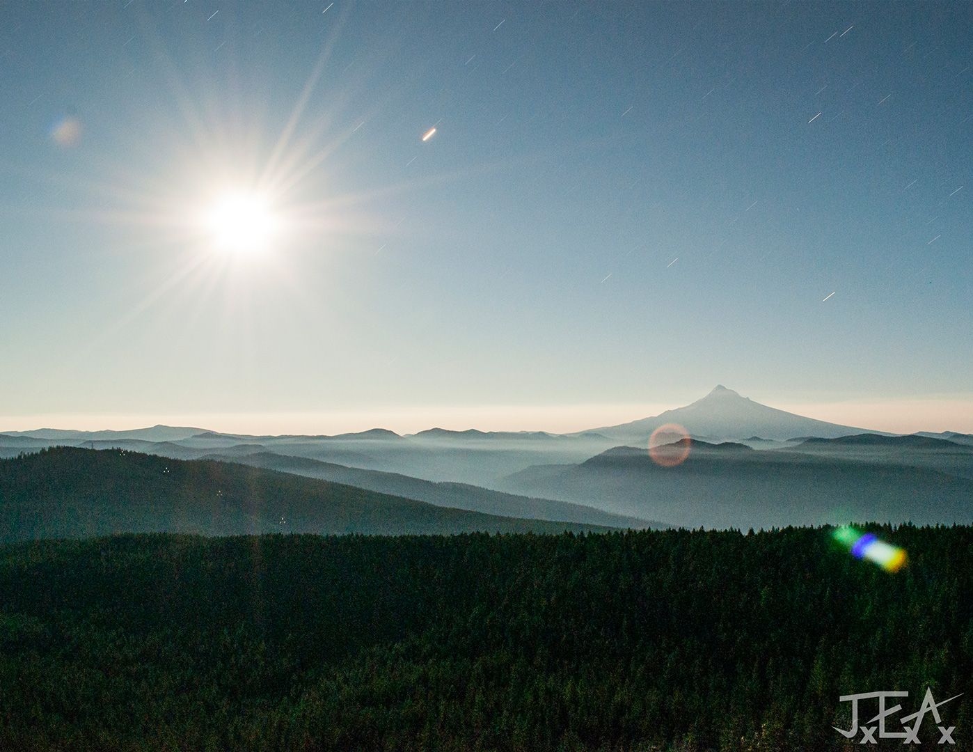 Mt Hood and a bright shining moon both sit in the distance over foggy forested hills.