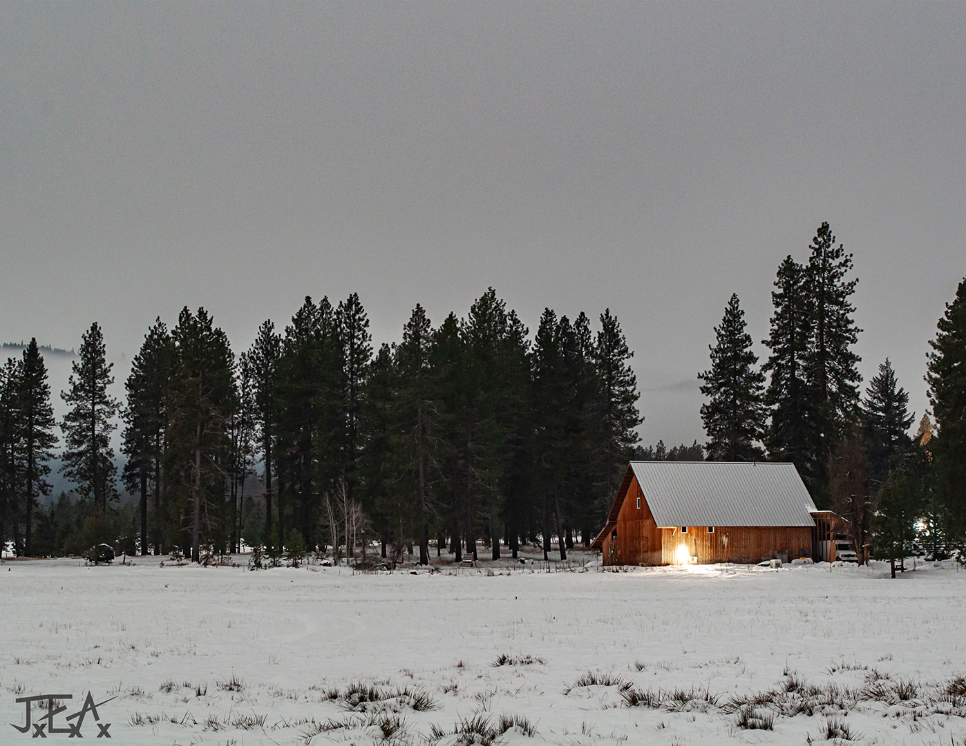 A farmhouse with light in the window sits in a snowwy field backed by evergreens on a winter night.