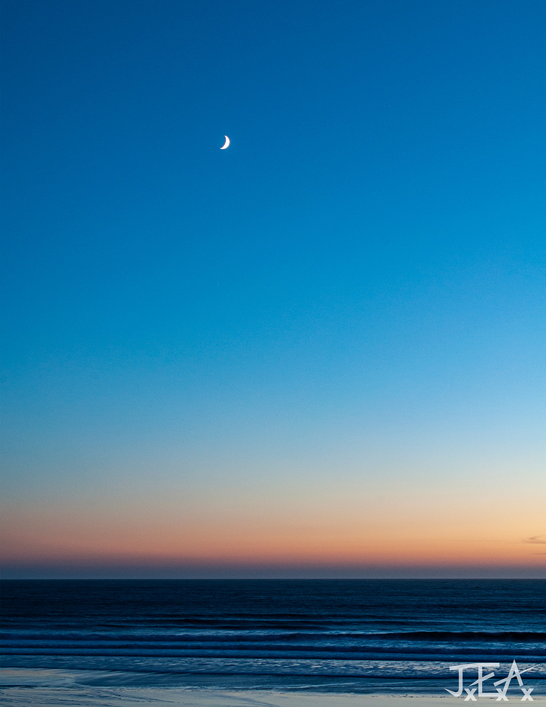 A wide shot of the Pacific Ocean bay at sunset, with a fingernail crescent moon hanging high above the horizon.